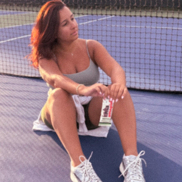 Athletic woman resting on tennis court holding an Attagirl Chocolate Sea Salt protein bar post-workout